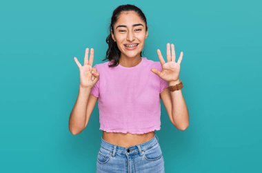 Diş teli takan, günlük kıyafetler giyen İspanyol genç kız sekiz numaralı parmaklarıyla gülümseyerek kendinden emin ve mutlu bir şekilde onu işaret ediyor.. 