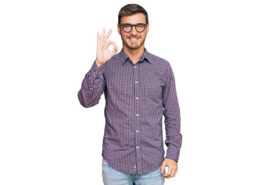 Yakışıklı beyaz bir adam günlük kıyafetler ve gözlük takıyor gülümsüyor ve el ve parmaklarıyla tamam işareti yapıyor. başarılı ifade. 