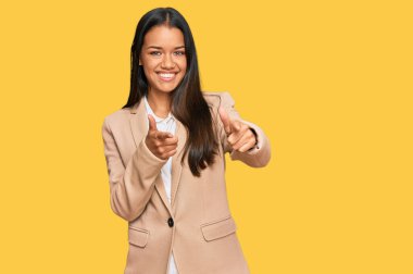 İş ceketi giyen güzel İspanyol kadın mutlu ve komik yüzlü parmaklarını kameraya doğrultuyor. iyi enerji ve titreşimler. 