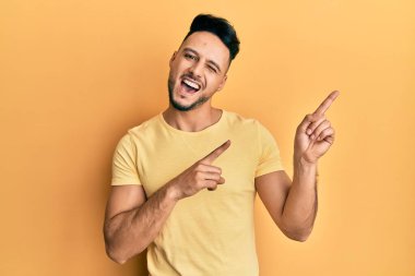 Gündelik kıyafetler giyen genç Arap adam gülümseyip kameraya bakıyor ve iki eliyle ve parmaklarıyla kenarı işaret ediyor.. 