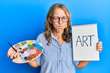 Orta yaşlı sarışın kadın boyacı paleti tutuyor ve defter şişiren komik yüzlü yanakları var. Ağzı havayla şişer, havayı yakalar.. 