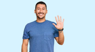 Dövmeleri olan yakışıklı bir adam. Günlük giysiler giyiyor. Beş numarayı gösteriyor ve gülümsüyor.. 