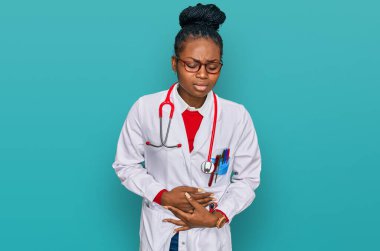 Genç Afro-Amerikan kadın doktor üniforması giyiyor ve steteskop kullanıyor midesi üstünde çünkü hazımsızlık, acı veren hastalıklar kendini iyi hissetmiyor. Ağrı kavramı. 