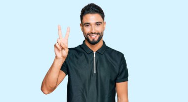 Sakallı genç adam spor giysisi giyiyor ve iki numaralı parmaklarıyla işaret ederken kendinden emin ve mutlu gülümsüyor.. 