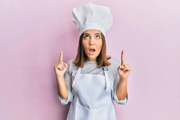 身穿职业厨师制服和帽子的年轻漂亮女人惊奇地抬起头 用手指和胳膊指指着 — 图库照片