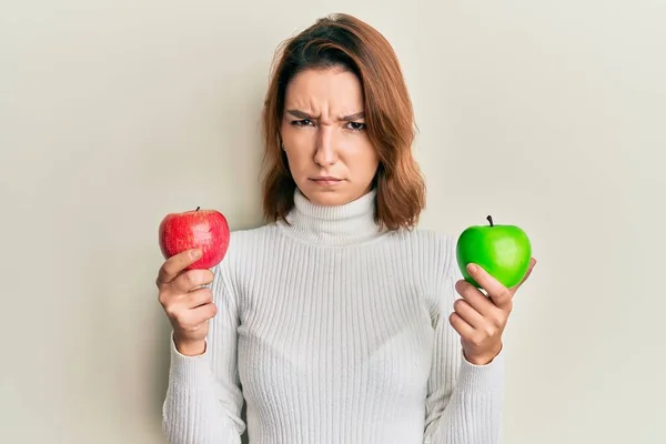 年轻的高加索女人 手里拿着红绿相间的苹果怀疑和紧张 皱着眉头因为问题而烦恼 消极的人 — 图库照片