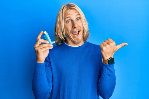 Caucasian young man with long hair holding medical asthma inhaler pointing thumb up to the side smiling happy with open mouth