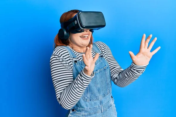 Young redhead woman scared using 3d virtual glasses standing over isolated blue background.