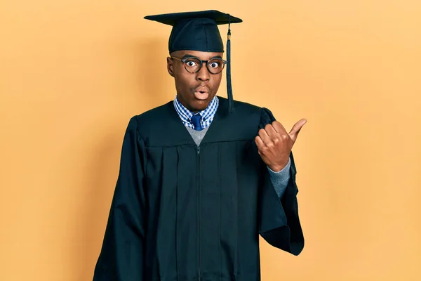 Młody Afroamerykanin Noszący Czapkę Dyplomową Szatę Ceremonialną Zaskoczony Wskazując Palcem — Zdjęcie stockowe