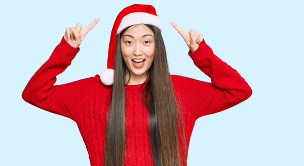 若いです中国人女性身に着けているクリスマスの帽子笑顔と驚きと指摘指と上げ腕 — ストック写真