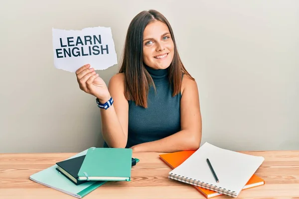 Young caucasian woman holding learn english paper while studying looking positive and happy standing and smiling with a confident smile showing teeth