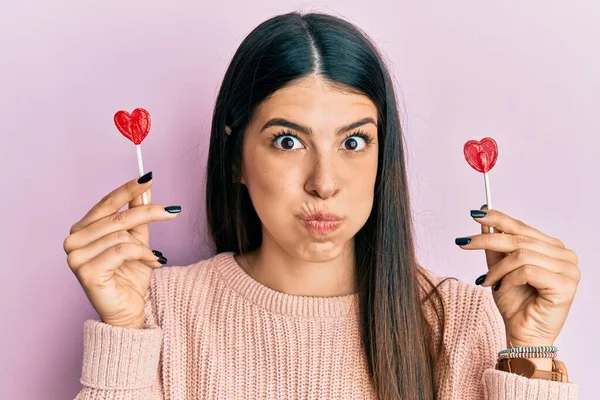 Young Hispanic Woman Holding Heart Lollipops Puffing Cheeks Funny Face — 스톡 사진