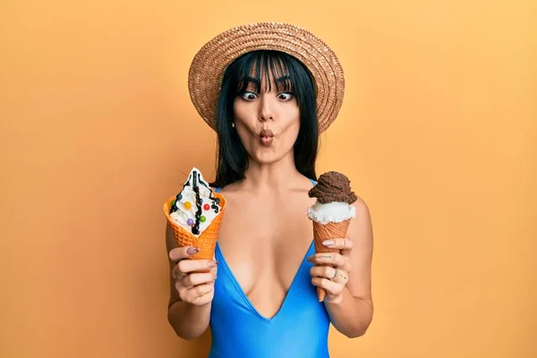 Young brunette woman with bangs wearing swimwear holding two ice cream cones making fish face with mouth and squinting eyes, crazy and comical.