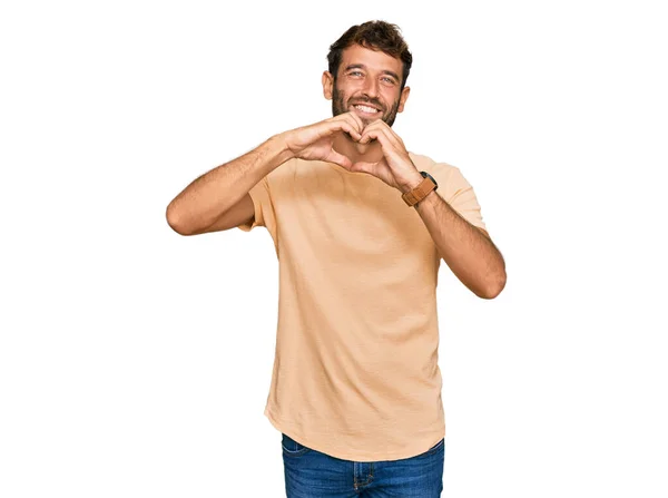 ひげを生やしたハンサムな若い男は 手でハートシンボルの形をする愛に笑みを浮かべてカジュアルなTシャツを着ています ロマンチックなコンセプト — ストック写真