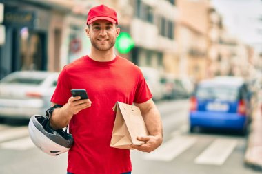Beyaz atlı genç teslimatçı akıllı telefon kullanıyor ve şehirde kağıt torba taşıyor..