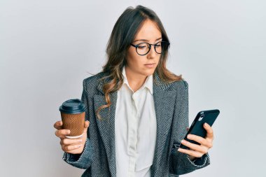 Genç esmer kadın akıllı telefon kullanıyor ve bir fincan kahve içiyor. Yüzünde ciddi bir ifade var. Basit ve doğal bir şekilde kameraya bakıyorsun.. 