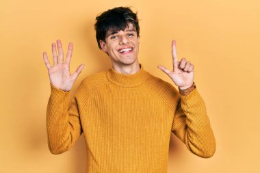 Yakışıklı hippi genç adam gündelik sarı süveter giyiyor ve yedinci parmaklarını gösterip gülümserken kendinden emin ve mutlu. 