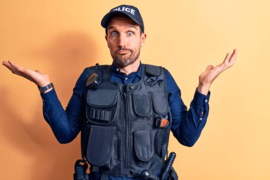 Young handsome policeman wearing police uniform and bulletprof over yellow background clueless and confused with open arms, no idea and doubtful face.