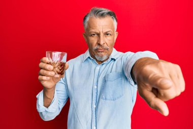 Middle age grey-haired man drinking glass of whisky pointing with finger to the camera and to you, confident gesture looking serious 