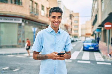 Genç Latin adam mutlu bir şekilde gülümsüyor ve şehirde akıllı telefon kullanıyor..