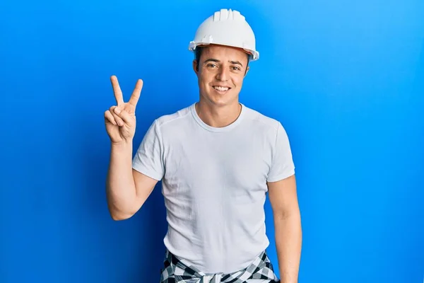 英俊的年轻人 身穿建筑工人制服 头戴硬礼帽 手握二指 面带微笑 自信而快乐 — 图库照片