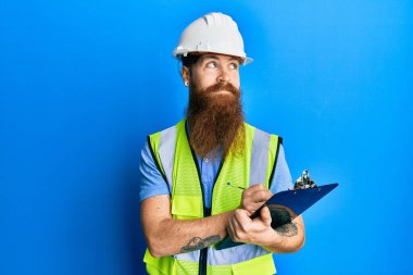Uzun sakallı, güvenlik kaskı takan, elinde pano tutan, gülümseyen ve kafasını çeviren kızıl saçlı bir adam.. 