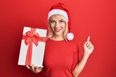 Genç sarışın kız Noel şapkası takıyor ve elinde bir hediye tutuyor. Gülümsüyor, mutlu bir şekilde işaret ediyor. 