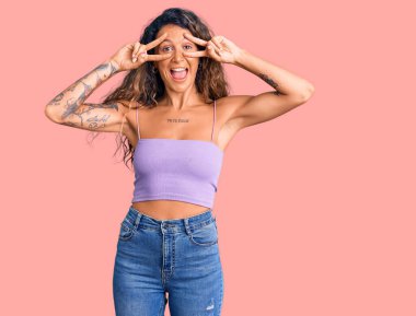 Dövmeli genç İspanyol kadın, gündelik kıyafetler giyiyor, barış sembolü yapıyor. Yüzünde parmakları var, gülümsüyor, zafer işareti yapıyor. 