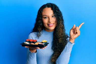 Young latin woman showing a plate of sushi smiling happy pointing with hand and finger to the side 