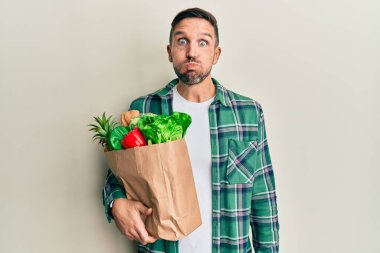 Handsome man with beard holding paper bag with groceries puffing cheeks with funny face. mouth inflated with air, crazy expression. 