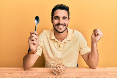 Genç İspanyol adam sağlıklı tahıllı yiyecekler yiyor masaya oturmuş gururla bağırıyor, zaferi kutluyor ve kollarını kaldırarak çok heyecanlanıyor. 