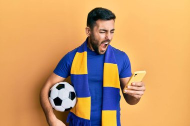 Genç İspanyol futbolcu holigan elinde futbol topu tutarken akıllı telefon kullanıyor kızgın ve öfkeli çığlıklar atıyor öfkeyle bağırıyor. öfke ve agresif konsept. 