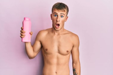 Young caucasian man shirtless holding shampoo bottle scared and amazed with open mouth for surprise, disbelief face 