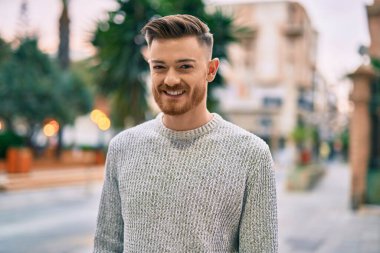 Genç beyaz adam gülümseyerek şehirde mutlu mesut duruyor..