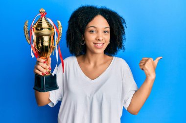 Young african american girl holding winner trophy pointing thumb up to the side smiling happy with open mouth 