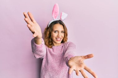 Paskalya tavşanı kulaklı beyaz bir kadın kucak açmak için kollarını açmış kameraya bakıyor. Mutluluğu kucaklayan neşeli bir ifade.. 