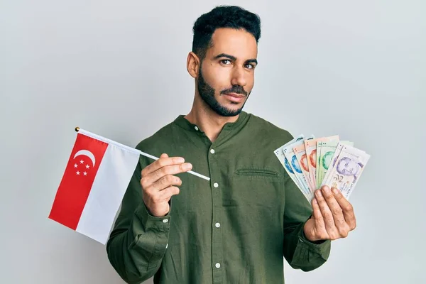 Young hispanic man holding singapore flag and dollars smiling looking to the side and staring away thinking.