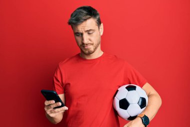 Futbol topu tutan yakışıklı beyaz adam akıllı telefondan habersiz ve kafası karışık bir ifadeye bakıyor. şüphe kavramı. 