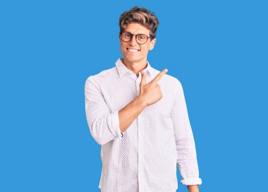 İş kıyafetleri giymiş, gözlüklü, neşeli, yüzünde gülümseme olan genç ve yakışıklı bir adam mutlu ve doğal bir ifadeyle elini ve parmağını kaldırıyor. 