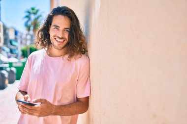 Duvara dayanmış akıllı telefon kullanan, mutlu gülümseyen genç İspanyol adam.