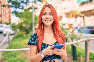 Genç kızıl saçlı kız şehirde akıllı telefon kullanarak mutlu bir şekilde gülümsüyor..