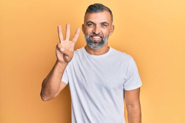 Orta yaşlı, beyaz tişört giyen yakışıklı bir adam üç numarayı gösterip işaret ederken kendinden emin ve mutlu bir şekilde gülümsüyor.. 