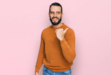 Sakallı genç adam, sıradan bir kazak giyiyor, gülümsüyor, gülümsüyor ve başparmağıyla yan tarafa bakıyor.. 