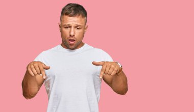 Yakışıklı, beyaz tişört giyen, reklamını gösteren parmaklarıyla aşağıya bakan kaslı adam, şaşırmış yüz ve ağzı açık. 