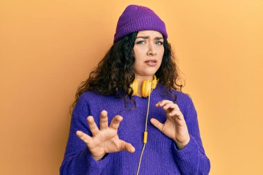 Young brunette woman with curly hair listening to music using headphones disgusted expression, displeased and fearful doing disgust face because aversion reaction. with hands raised 