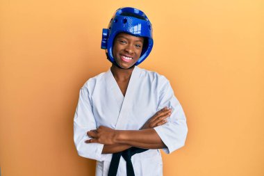 Tekvando kimonosu ve koruyucu başlık takan, kollarını kavuşturup kameraya gülümseyen genç Afrikalı Amerikalı kız. pozitif kişi. 