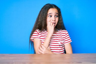 Gündelik kıyafetler giyen tatlı İspanyol kız masada oturmuş gergin ve elleri ağızda, tırnaklarını yiyormuş gibi görünüyor. anksiyete. 