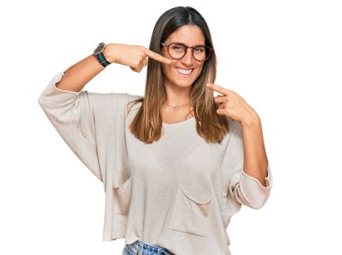 Gündelik kıyafetler ve gözlük takan genç bir kadın neşeli bir şekilde gülümseyip parmaklarını ve ağzını işaret ediyor. diş sağlığı kavramı. 