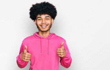Afro saçlı, günlük pembe kazak giyen, başarı işareti yapan eli olumlu hareket eden, gülümseyen ve mutlu genç Afro-Amerikalı bir adam. Neşeli ifade ve kazanan jest. 