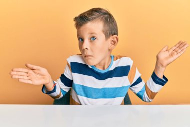 Gündelik giysiler giyen sevimli beyaz çocuk masanın üzerinde elleri ve kolları havada, kafası karışmış bir şekilde oturuyor. şüphe kavramı. 